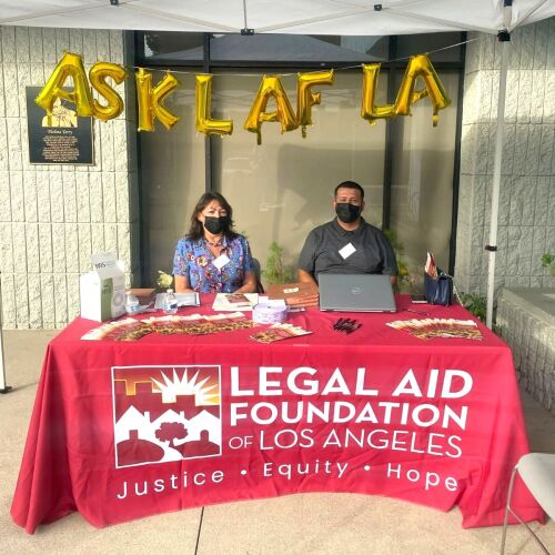 Two people sit at a table with a red LAFLA tablecloth beneath gold balloons reading ASK LAFLA.