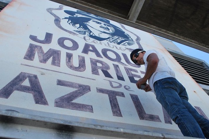 A photo taken from ground level shows artist Willie Herron standing elevated in front of the Joaquin Murieta de Aztlan mural. He is looking down at the camera.